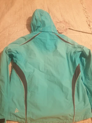 Turkos vindjacka från Craft, stl S - Turkos vindjacka från Craft med svarta och vita detaljer. Jackan har huva, dragkedja och meshfoder på insidan för extra ventilation. Snygg sportig look med smidiga fickor och justerbar nederkant. Perfekt för dig som gillar att vara aktiv utomhus.