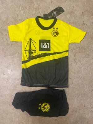 Borussia Dortmund fotbollskit barn - Snyggt fotbollskit för barn med Borussia Dortmunds gula och svarta färger. Tröjan har klubbens logga, sponsortryck och motiv framtill samt spelarnamn och nummer 5 på ryggen. Matchande svarta shorts med klubbmärke. Perfekt för unga fans!