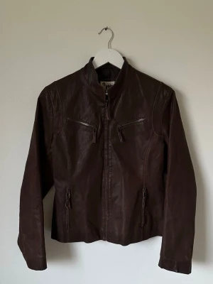 Brun skinnjacka från Joy - Snygg brun skinnjacka med dragkedja framtill och två zip-fickor på bröstet. Jackan har en stilren, rak passform och är helfodrad med polyester. Perfekt för dig som vill ha en klassisk look med edge. 7Y8