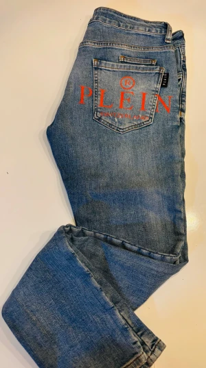 Philipp Plein ljusblå jeans med tryck - Ljusblå jeans från Philipp Plein med röd logotyp på bakfickan och slitna detaljer framtill. Jeansen har klassisk femficksdesign, silverfärgad knapp och små nitar vid fickorna. Fit och passformen slimfit, super straight cut, använda två gånger   Size 30 Lenght top/bottom Front 103 cm Back 108 cm Inner 82 cm  Nypris 4999kr 