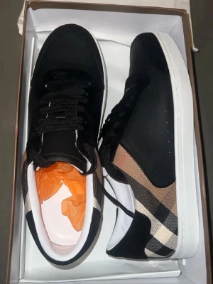 Burberry sneakers med klassiskt rutmönster - Snygga svarta sneakers från Burberry med den ikoniska beige och svarta rutan på sidorna. Skorna har vita sulor, svarta snören och detaljer i skinn och textil. Perfekta för dig som vill ha en stilren och lyxig look med en touch av brittisk klass.
