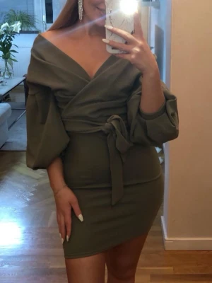 Olivgrön omlottklänning från Boohoo - Snygg olivgrön kort omlottklänning från Boohoo med puffiga långärmar och knyt i midjan. Klänningen har v-ringad hals 