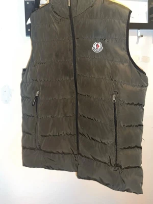 Moncler mörkgrön dunväst med dragkedja - Säljer en mörkgrön dunväst från Moncler med quiltad design och två fickor med dragkedja framtill. Västen har ett broderat Moncler-märke på bröstet och svart foder med Moncler-tryck på insidan. Perfekt för lager-på-lager under kyliga dagar.