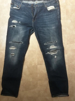 Premium Jeans blå slitna herrjeans - Blå jeans från Premium Jeans med snygga slitningar och hål på framsidan. Klassisk femficksmodell med raka ben och normal passform. Jeansen har en mörk tvätt och är tillverkade i jeansmaterial med kontrastsömmar. Perfekt för dig som gillar en avslappnad och trendig look.