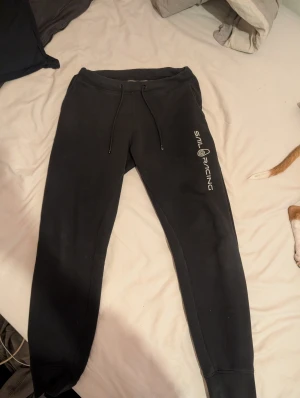 Svarta mjukisbyxor Sail Racing  - Svarta mjukisbyxor från Sail Racing i storlek S. Byxorna har snörning i midjan, en bakficka och logotypen tryckt längs benet. Sköna och stilrena med smal passform, perfekta för chill eller träning. Materialet är mjukt och känns som bomull eller polyester.