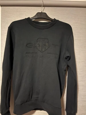 Svart sweatshirt från GANT, storlek S - Svart sweatshirt från GANT med broderad logga och text på bröstet. Tröjan har rund hals, ribbade muddar och är långärmad. Skön tröja som är perfekt för kyligare säsonger och passar till allt. Perfekt för dig som gillar stilren och klassisk design.🤩
