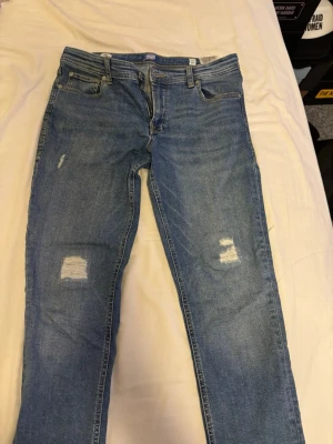Blå jeans från Jack & Jones, 164 - Säljer ett par blå jeans från Jack & Jones i storlek 164. De har klassisk femficksdesign, slitna detaljer på framsidan och en snygg läderpatch med logga bak. Jeansen har rak passform och är gjorda i jeansmaterial med gul kontrastsöm.