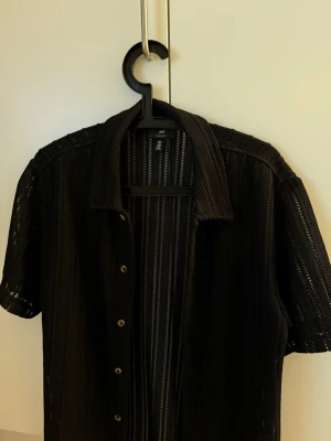 Svart stickad skjorta  - Svart stickad skjorta från H&M med kort ärm och knappar framtill. Skjortan har ett genomgående hålmönster i vertikala ränder som ger en luftig och cool känsla. Perfekt att slänga över ett linne eller t-shirt för en avslappnad vibe.