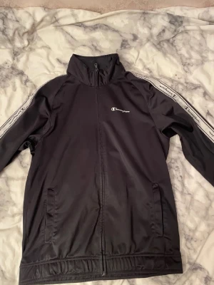 Champion tracksuit set - Bilderna visar ett svart Champion tracksuit-set med jacka och byxor, men inga skor finns med.