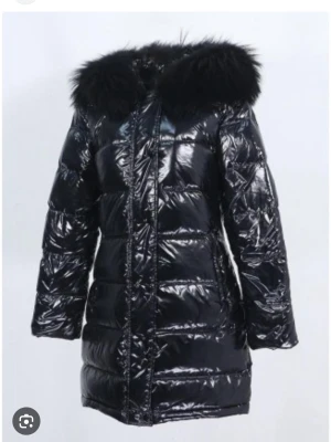 Svart glansig pufferjacka med päls - Säljer en svart, glansig pufferjacka med fluffig svart päls på huvan. Jackan har lång passform, dragkedja framtill och quiltade sektioner. Perfekt för kalla dagar när du vill vara både varm och snygg.