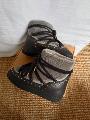 Inuikki skor - Säljer ett par grå boots med mjuk päls på insidan och utsidan, svarta snören med gulddetaljer och svarta gummisulor. Skorna har dekorativa nitar runt hela kanten och är perfekta för kalla dagar. Snygg och unik design som sticker ut.