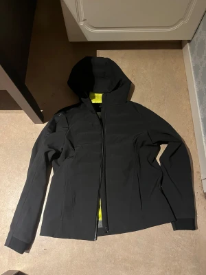 Svart vindjacka från Sail Racing - Svart vindjacka från Sail Racing med huva och hel dragkedja framtill. Jackan har en stilren design med diskreta logodetaljer på ärmarna och insida i gul färg. Materialet är syntet och jackan har långärmad passform, perfekt för blåsiga dagar. Den är M i damstorlek men XSS i herr storlek