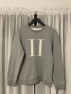 Grå sweatshirt från Les Deux - Snygg grå sweatshirt från Les Deux med stort vitt II-tryck på bröstet. Tröjan har rund hals, ribbade muddar och är långärmad. Perfekt för chill dagar och enkel att matcha med jeans eller joggers.