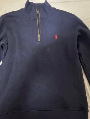Marinblå half zip tröja från Polo Ralph Lauren - Säljer en marinblå långärmad tröja från Polo Ralph Lauren med half zip och röd broderad logga på bröstet. Tröjan har en hög krage och är gjord i mjuk bomull, perfekt för chill dagar. Passar dig som gillar klassisk stil med en sportig touch. Helt ny passar inte. Priset kan diskuteras vid snabb affär.