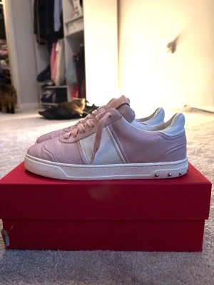 Valentino Garavani rosa sneakers - Säljer ett par snygga Valentino Garavani sneakers i ljusrosa och vitt skinn med klassiska nitar på sulkanten. Skorna har rund tå, platt sula och snörning. Märkeslogga på plösen och yttersulan. Perfekta för dig som gillar exklusiva detaljer och stilren design. Gjort för tjejer men funkar säkert som unisex. Man får med box snören och kvitto🫰🏻💐❤️