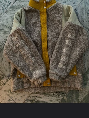 Adidas teddyjacka med broderi, S - Säljer en teddyjacka från Adidas med beige och gula detaljer. Jackan har broderad text 'STRIPES' och 'THREE' på ärmarna, hög krage och tryckknappar. Materialet är mjuk fleece och polyester, perfekt för kalla dagar. Se bilder för storlek och skick. Fick i gåva så osäker om det är dam eller herr. Men funkar för mig som vanligtvis har small i herr fast då blir ärmarna bara en aning lite för korta. Skulle gissa på att det är unisex.