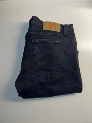 Nudie Jeans - Säljer ett par svarta skinny jeans från Nudie Jeans. Jeansen har en mörk tvätt och Nudie-logga på bakfickan. Materialet är stretchig bomull med snygg passform och smala ben. Midja 39cm, längd 102cm. Går ej att se storleken på lappen men L storleken är L32 midjan bedöms till W30 men utgå från måtten. JAG SKICKAR EJ FLER BILDER NÄR JAG BÄR JEANSEN! för storleksguide hänvisar jag till måtten och för bättre inblick i passformen rekommenderar jag att googla på modellnamnet. kolla måtten noggrant :)s5
