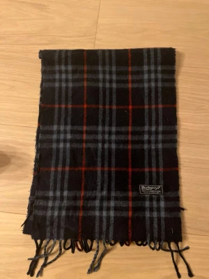 Rutig ullväst från Burberrys - Säljer en klassisk rutig väst från Burberrys i mörkblå och röda toner. Västen är tillverkad i 100% lammull och har ett snyggt mönster med fransar nedtill. Perfekt för dig som gillar tidlös design och vill ha något som sticker ut.