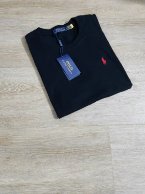 Svart t-shirt från Polo Ralph Lauren - Svart t-shirt från Polo Ralph Lauren i storlek L. Klassisk design med rund hals och korta ärmar, tillverkad i mjuk bomull. På bröstet finns den ikoniska röda Polo-loggan broderad. Perfekt för en clean och stilren look.