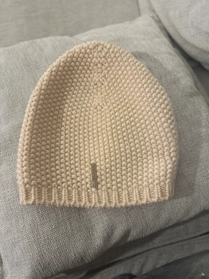 Prada Beige stickad mössa  - Snygg beige stickad mössa från Prada, tillverkad i 100% virgin ull. Mössan har ett grovt stickat mönster och en liten metallbricka med Prada-loggan framtill. Perfekt för dig som gillar exklusiva accessoarer med enkel design.