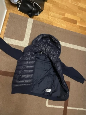 Mörkblå pufferjacka från Maggiore - Säljer en mörkblå pufferjacka från Maggiore med huva och dragkedja. Jackan har quiltad framsida och rygg i nylon, samt stickade ärmar med Maggiore-logga på ena armen. Materialet är nylon och polyester, vilket gör den både lätt och varm. Perfekt för kyliga dagar!