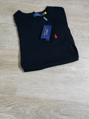 Svart t-shirt från Polo Ralph Lauren - Svart t-shirt från Polo Ralph Lauren med klassisk rund hals och korta ärmar. T-shirten har en röd broderad logga på bröstet och är gjord i mjuk bomull. Perfekt för dig som gillar stilrena och tidlösa plagg med en touch av lyx.