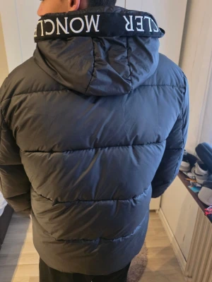 Svart Moncler pufferjacka med huva - Svart pufferjacka från Moncler med stor huva och logotyp på insidan. Jackan har dragkedja framtill, två sidofickor och en clean, stilren look. Materialet är syntet och dun, perfekt för kalla dagar. Moncler-etikett på insidan och på huvan för extra vibe. 