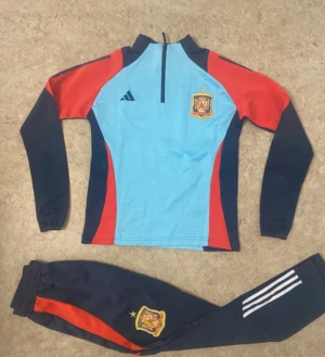 Spaniens Adidas fotbollsträningsoverall - Säljer en officiell träningsoverall från Spaniens landslag, Adidas. Setet består av en långärmad tröja i ljusblått, rött och mörkblått med halvlång dragkedja och broderad landslagslogga, samt matchande byxor med röda detaljer och vita ränder. Perfekt för fotbollsträning.