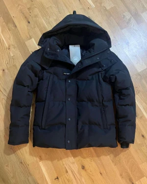 Svart dunjacka från Canada Goose - Svart dunjacka från Canada Goose med stor huva och diskret logopatch på ärmen. Jackan har dragkedja och knappar framtill, samt två sidofickor. Den är tjockt fodrad och har en clean, stilren look som funkar perfekt när det är kallt ute.