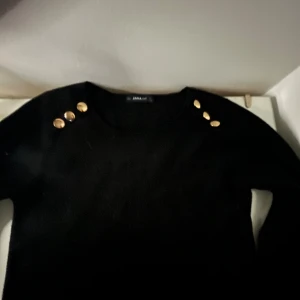 Svart stickad tröja med guldknappar Zara - Svart stickad tröja från Zara med rund halsringning och snygga guldknappar på axlarna. Tröjan har lång ärm och en klassisk, stilren look som passar perfekt till jeans eller kjol. Materialet är mjukt och bekvämt för höst och vinter.