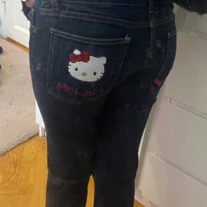 Hello Kitty jeans med broderad bakficka - Mörkblå jeans från Hello Kitty med broderad Hello Kitty-figur och röd rosett på bakfickan. Klassisk femficksmodell med raka ben och kontrastsömmar. Snygg detalj med Hello Kitty-märke och knapp framtill. Perfekt för dig som gillar unika plagg! Äkta Sanrio och säljs inte längre, priset kan diskuteras