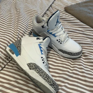 Nike Air Jordan 3 Vit/Blå Sneakers - Nike Air Jordan 3 sneakers i vitt skinn med blå detaljer och ikoniskt grått elephant-mönster på tå och häl. Rund tå, snörning och platt sula. Snygg retrostil med Jumpman-logga på plösen. Perfekt för dig som gillar klassiska och sportiga sneakers.