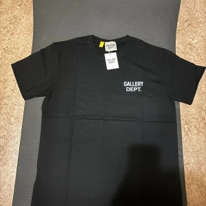 Svart t-shirt från Gallery Dept. - Svart t-shirt från Gallery Dept. med vit logga tryckt på bröstet och stor text på ryggen. Klassisk rund hals och korta ärmar. Tillverkad i mjuk bomull för en skön känsla. Perfekt för dig som gillar streetwear och enkel stil.