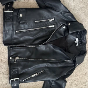 Svart skinnjacka från Morgan - Snygg svart skinnjacka från Morgan med flera silvriga dragkedjor, bälte i midjan och klassisk krage. Jackan har cool bikerstil och detaljer som tryckknappar och fickor med dragkedja. Perfekt för dig som vill ha en edgy look.