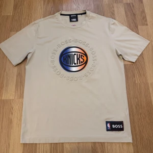 Beige Knicks t-shirt från BOSS x NBA - Beige t-shirt från BOSS x NBA med New York Knicks-logga i blått och orange framtill. Rund hals, korta ärmar och patch med NBA och BOSS vid nederkanten. Skön bomullskvalitet och clean design, perfekt för dig som gillar basket och streetstyle.