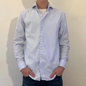 Ljusblå skjorta från Eton slim fit - Snygg ljusblå skjorta från Eton i slim fit-modell. Skjortan har klassisk krage, vita knappar och diskret mönstrad insida på kragen. Tillverkad i bomull. Modellen är 180 cm lång.