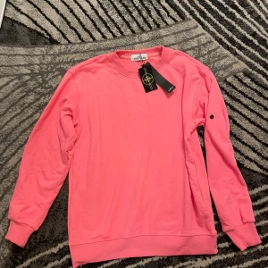 Rosa sweatshirt från Stone Island - Säljer en snygg rosa sweatshirt från Stone Island med klassisk rund hals och ribbade muddar. Tröjan har svarta knappar på ärmen och den ikoniska Stone Island-taggen. Tillverkad i mjuk bomull, perfekt för dig som gillar streetwear och vill sticka ut. Passar M.