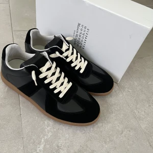 Maison Margiela svarta sneakers - Maison Margiela sneakers i svart skinn och mocka med vita snören och klassisk gummisula. Snygg minimalistisk design med rund tå och diskreta detaljer. Perfekta för dig som gillar stilrena och exklusiva sneakers.