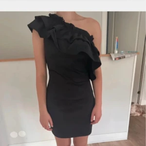 Svart one shoulder klänning med volang - Supersnygg svart kortklänning med one shoulder och stora volanger över axeln. Klänningen har en tight passform och är axelbandslös på ena sidan, vilket ger en cool och trendig look. Perfekt för dig som vill sticka ut med en unik siluett.