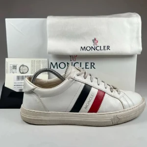 Moncler vita sneakers med röd och blå detalj - Snygga vita sneakers från Moncler i skinn med klassiska röd och marinblå ränder på sidan och Moncler-logga. Modellen har rund tå, platt sula och snörning. Perfekt för dig som gillar stilrena och exklusiva sneakers med en sportig touch.