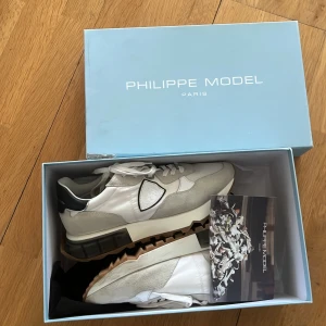 Philippe Model vita och beige sneakers - Snygga Philippe Model sneakers i vitt och beige med detaljer i svart. Skorna har en chunky platt sula i gummi med coolt mönster undertill, snörning och paneler i mocka och textil. Ikonisk sköldlogga på sidan och svart hälparti. Perfekt för dig som gillar stilrena och trendiga sneakers.