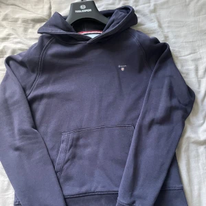Mörkblå hoodie från GANT - Säljer en stilren mörkblå hoodie från GANT med broderad logga på bröstet. Tröjan har huva, känguruficka och långa ärmar. Perfekt för dig som gillar klassisk och enkel streetstyle.