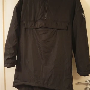 Svart jacka Urban Classics - Svart jacka från Urban Classics med huva och halv dragkedja framtill. Jackan har en stor ficka med lock på bröstet och ett snyggt rundat märke på ärmen. Materialet är lätt och syntetiskt, perfekt för blåsiga dagar. Passformen är loose och jackan är långärmad.