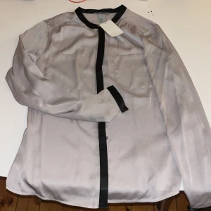Silvrig skjorta med svarta detaljer H&M - Snygg silvrig skjorta från H&M i storlek 36 med svarta kontrastdetaljer längs krage, knappslå och manschetter. Skjortan har två bröstfickor och är tillverkad i ett glansigt, mjukt material som ger en cool vibe. Perfekt för dig som gillar stilrena plagg med edge.