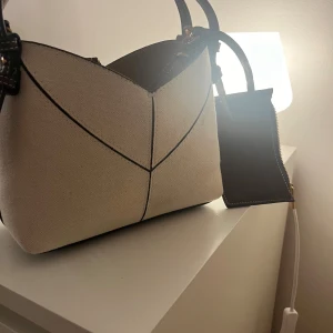 Beige axelväska med bruna detaljer - Snygg beige axelväska med bruna skinnremmar och matchande bruna detaljer. Väskan har en unik geometrisk form med svarta sömmar som ger en cool kontrast. Materialet är robust bomull och skinn, och den har en praktisk liten ficka med dragkedja på sidan.