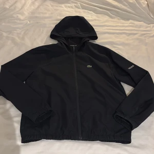 Mörkblå vindjacka från Lacoste  - Mörkblå vindjacka från Lacoste Sport med huva och hel dragkedja framtill. Jackan har elastiska muddar och nederkant, samt Lacoste-logga på bröstet och text på ärmen. Storlek XS