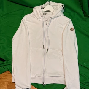 Vit hoodie från Moncler med dragkedja - Vit hoodie från Moncler med hel dragkedja, huva och snörning. Tricolore-rand på huvan och ärmslut, samt Moncler-logga på ärmen. Mjuk bomullskänsla och två fickor framtill. Perfekt för en clean och sportig look.Liknande modell som originalet!
