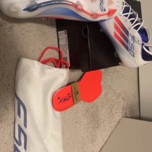Adidas F50 Elite fotbollsskor - Adidas F50 Elite fotbollsskor i vitt med röda och blå detaljer. Skorna har snörning, platt sula med dobbar och är tillverkade i syntetmaterial. Snygg, sportig design med F50-logga på sidan och medföljande skopåse och extra innersulor.