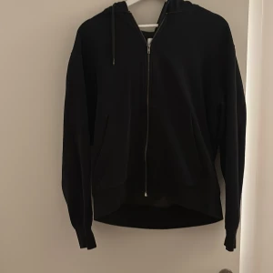 Svart hoodie från Jack & Jones - Svart hoodie från Jack & Jones med dragkedja framtill och luva med snörning. Mjuk fleece på insidan och två fickor. Perfekt för kyliga dagar och enkel att matcha med jeans eller joggers.