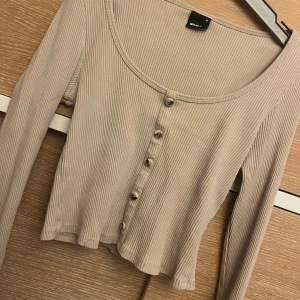 Beige ribbad topp med knappar Gina Tricot - Säljer en beige ribbad topp från Gina Tricot med lång ärm och rund halsringning. Toppen har snygga knappar framtill och är croppad i modellen. Perfekt att styla med jeans eller kjol för en chill look.